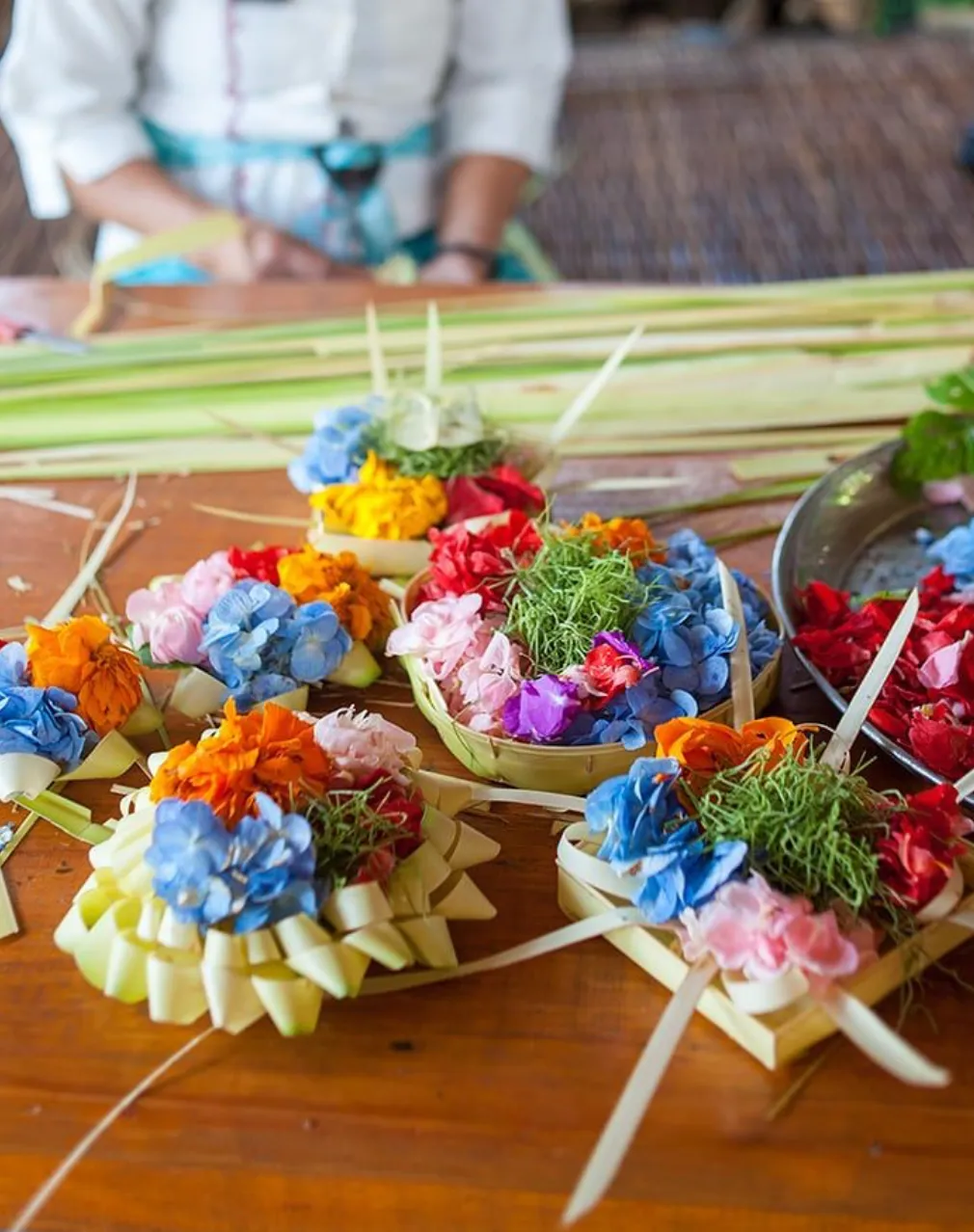 Workshop for making Balinese Canang Sari offerings, showing participants crafting small colorful floral arrangements used in daily Hindu rituals, set in a tropical setting.
