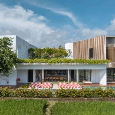 MandalaOasis_Bali_02_Exteriors_11zon A luxurious tropical villa named Mandala Oasis Villa, featuring an infinity pool surrounded by palm trees, lush gardens, and modern open-air architecture under a bright blue sky.