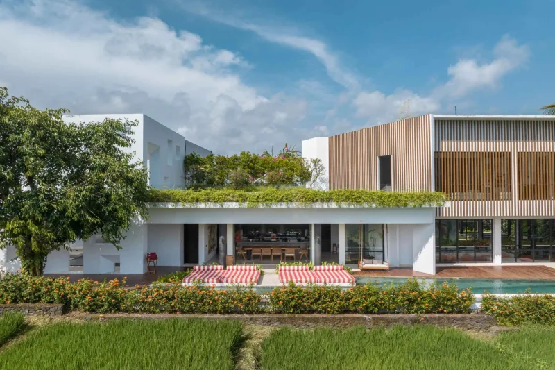 A luxurious tropical villa named Mandala Oasis Villa, featuring an infinity pool surrounded by palm trees, lush gardens, and modern open-air architecture under a bright blue sky.