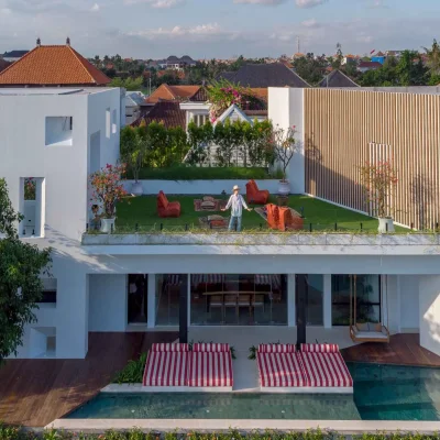 MandalaOasis_Bali_04_Exteriors_11zon A luxurious tropical villa named Mandala Oasis Villa, featuring an infinity pool surrounded by palm trees, lush gardens, and modern open-air architecture under a bright blue sky.