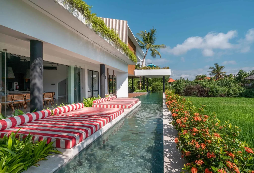 A luxurious tropical villa named Mandala Oasis Villa, featuring an infinity pool surrounded by palm trees, lush gardens, and modern open-air architecture under a bright blue sky.